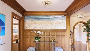 A reception area with a wooden reception desk, two patterned ottomans, a chandelier, and a large wall mural depicting a beach scene. - Channel Road Inn, A Four Sisters Inn (Santa Monica)