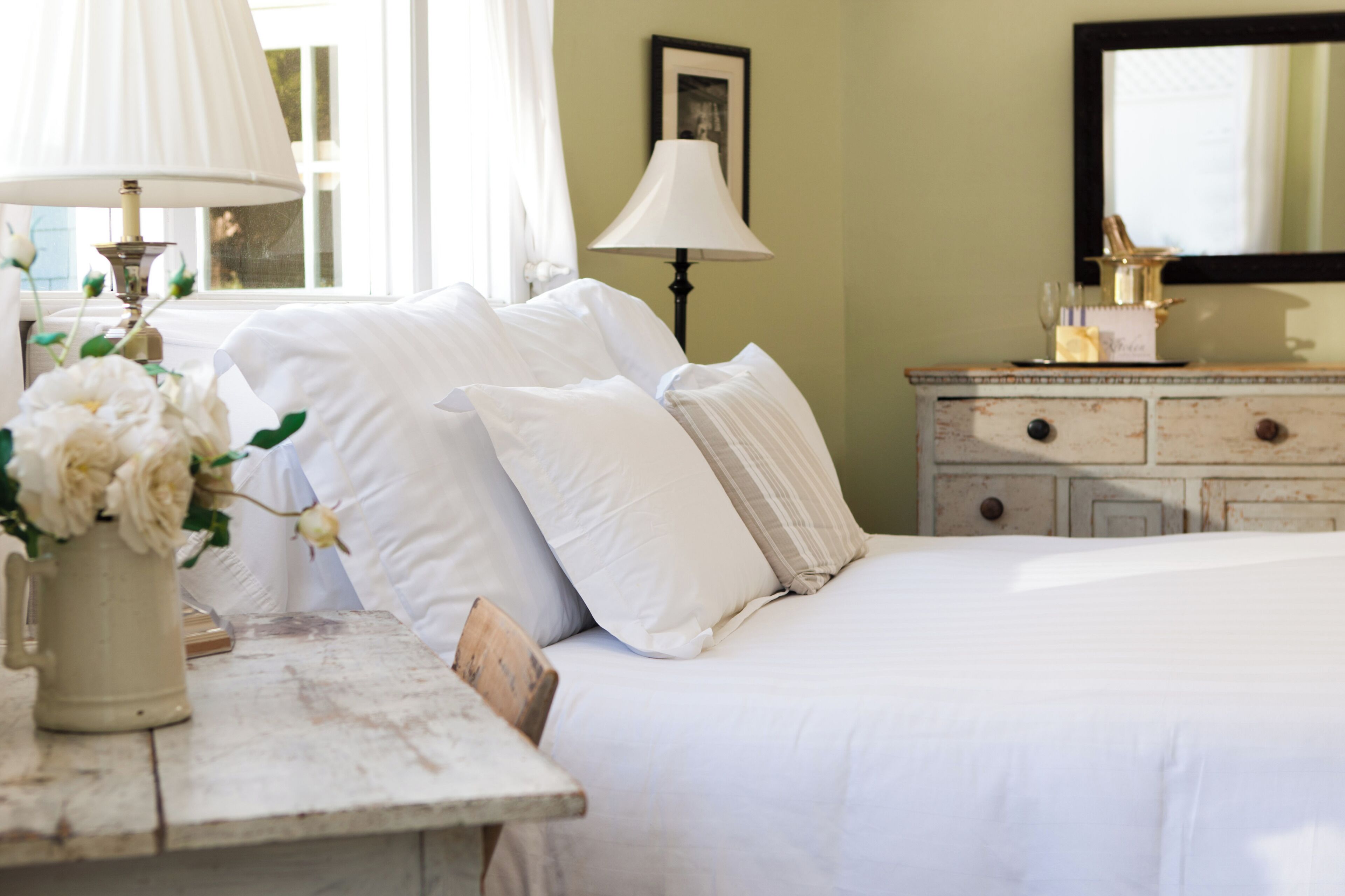 A neatly made bed with white linens and pillows, a wooden nightstand with a vase of flowers, and a dresser with drawers.