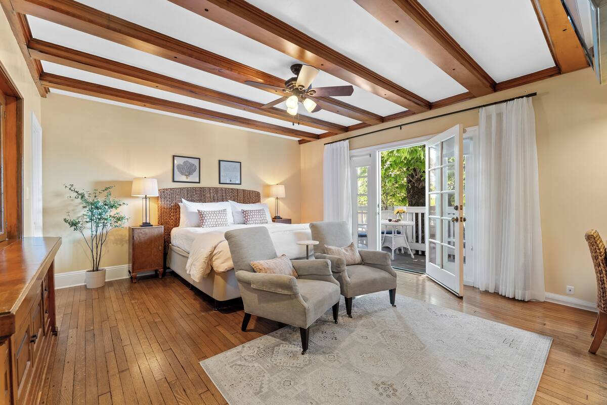 A spacious bedroom with a large bed, two armchairs, a wooden ceiling, and a view of the outdoors.