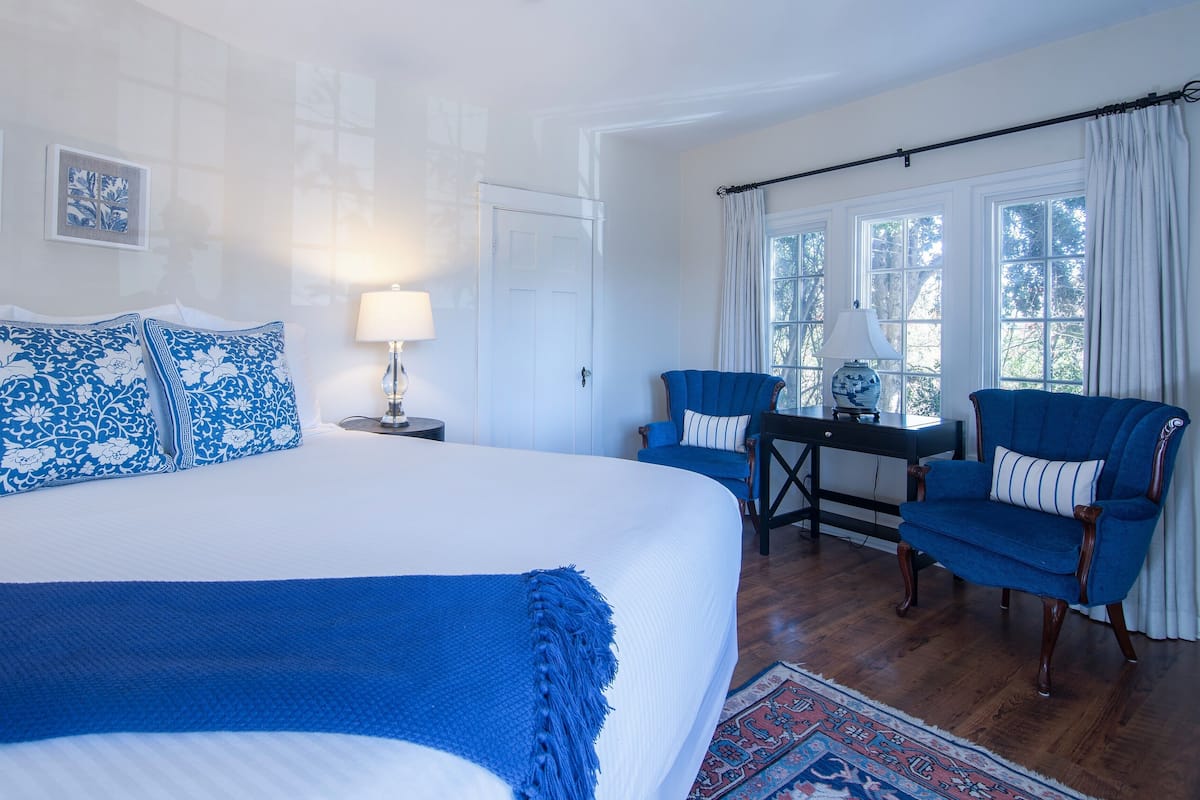 A bedroom with a large bed, two blue armchairs, a small table with a lamp, and a view of trees through the window.