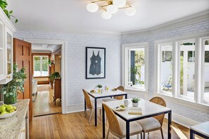 A dining area with a wooden floor, white walls, and large windows. There is a framed picture on the wall and a table set with plates and a pitcher.