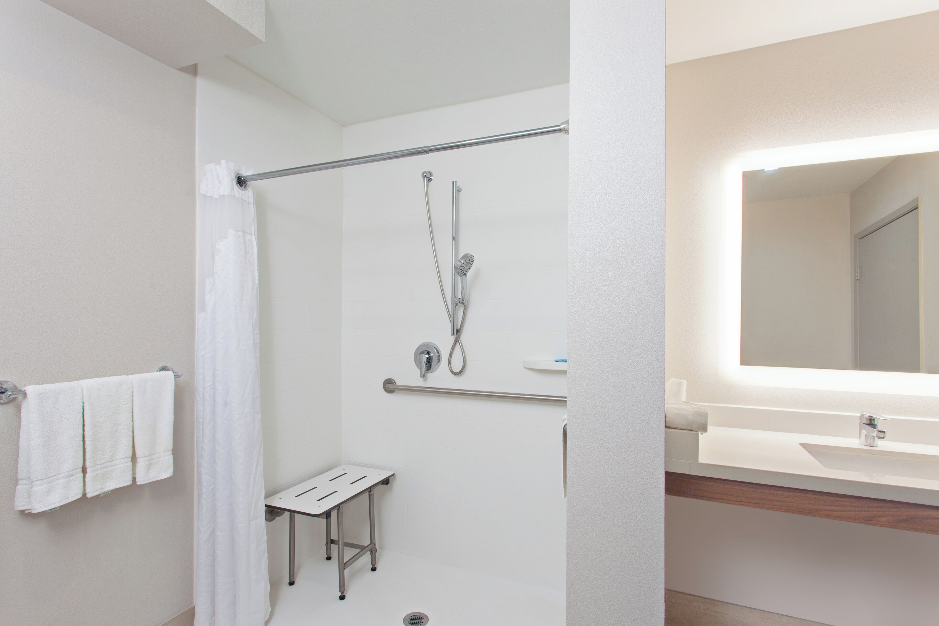 Une salle de bain avec une douche, un petit tabouret, un porte-serviettes et un lavabo avec un miroir.