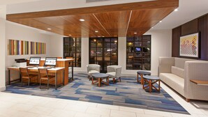 A modern lobby with a wooden ceiling, a seating area with sofas and chairs, a reception desk with computers, and large windows with city views.