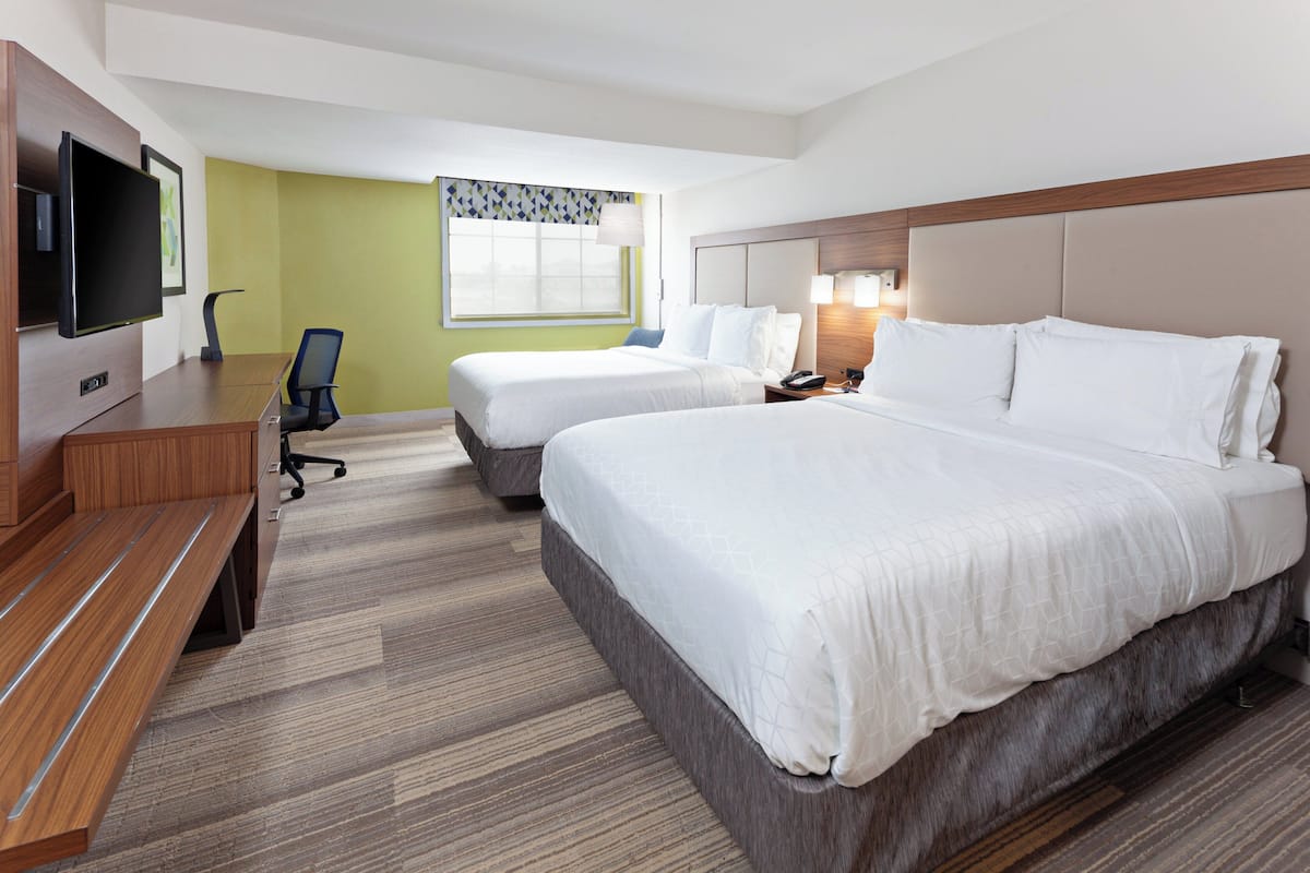 Une chambre d’hôtel avec deux lits, un bureau doté d’un téléviseur à écran plat, une chaise, une fenêtre avec un rideau à motifs et une lampe fixée au mur.