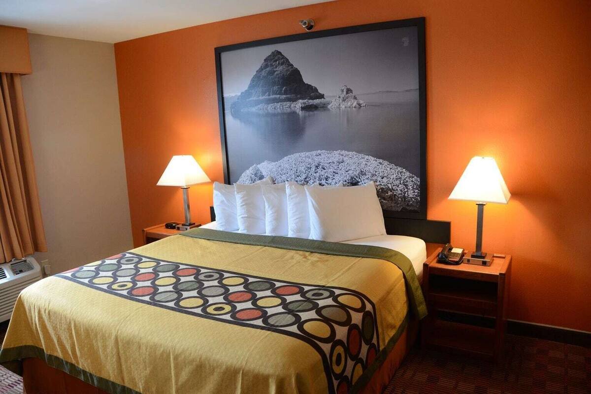 A hotel room with a bed, two bedside lamps, a large framed picture on the wall, and a window with curtains.
