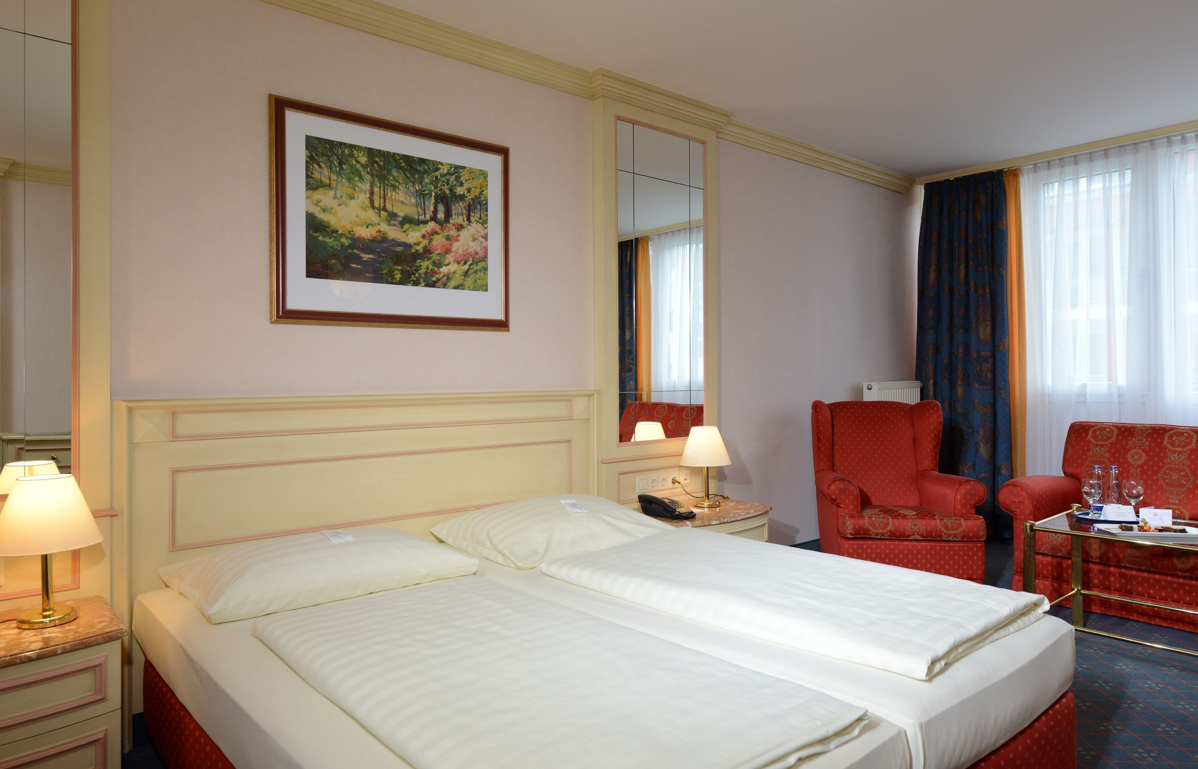 Une chambre d’hôtel avec deux lits, un fauteuil rouge, une petite table avec un verre d’eau et un tableau accroché au mur.