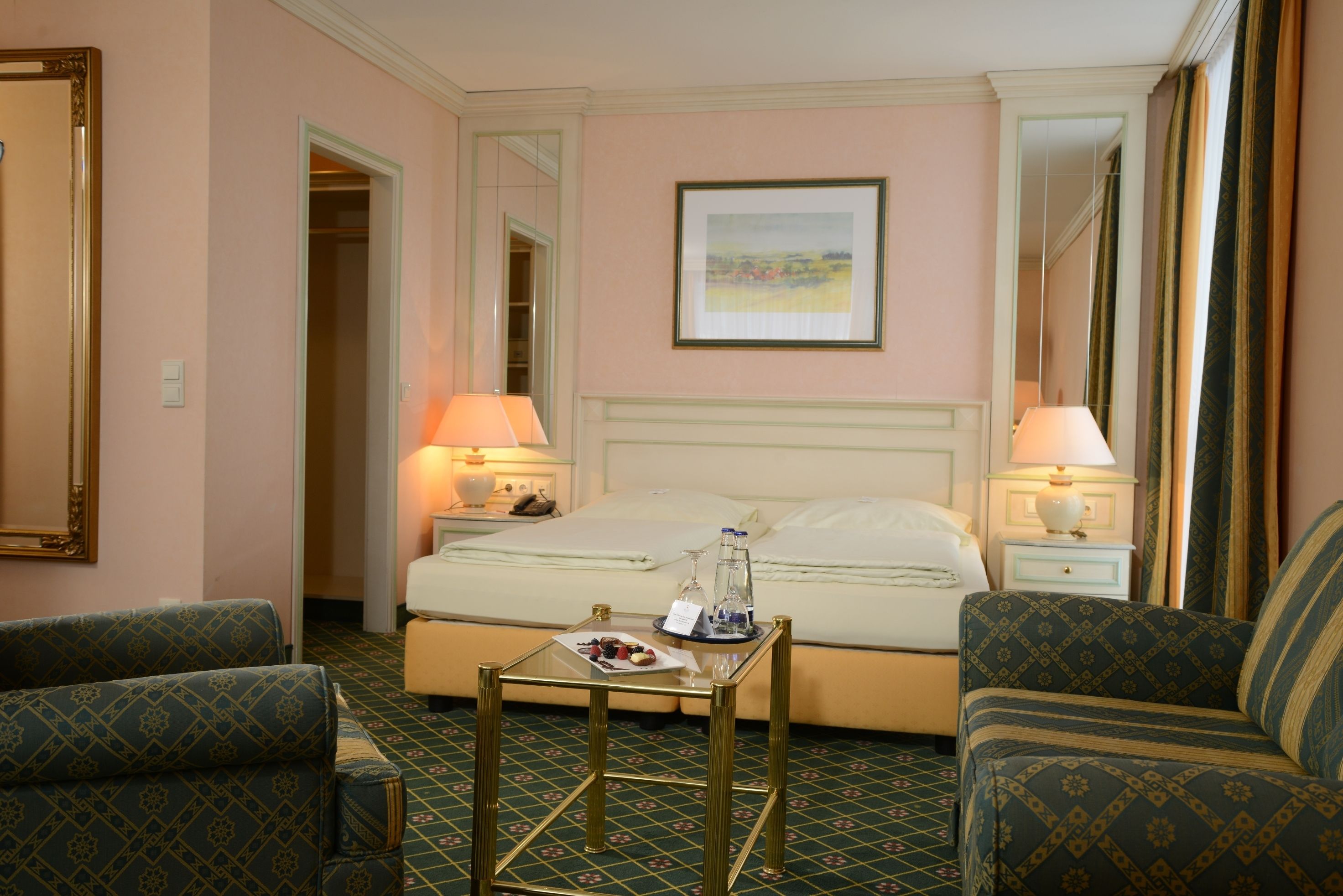 A hotel room with a bed, two bedside tables with lamps, a small table with a plate of food, and two patterned armchairs.