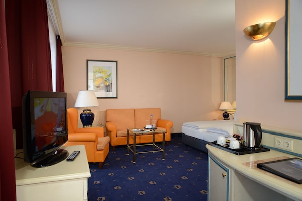 A hotel room with a television, orange sofa, a small table with a lamp, a bed, and a minibar. - The Monarch Hotel (Neustadt an der Donau)