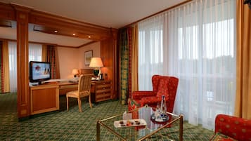 A hotel room with a television, desk, chair, sofa, and a glass coffee table with snacks.