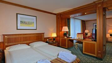 Ein Hotelzimmer mit zwei Betten, einem Schreibtisch und einem Stuhl. An der Wand hängt ein gerahmtes Bild und es gibt ein Fenster mit Vorhängen.