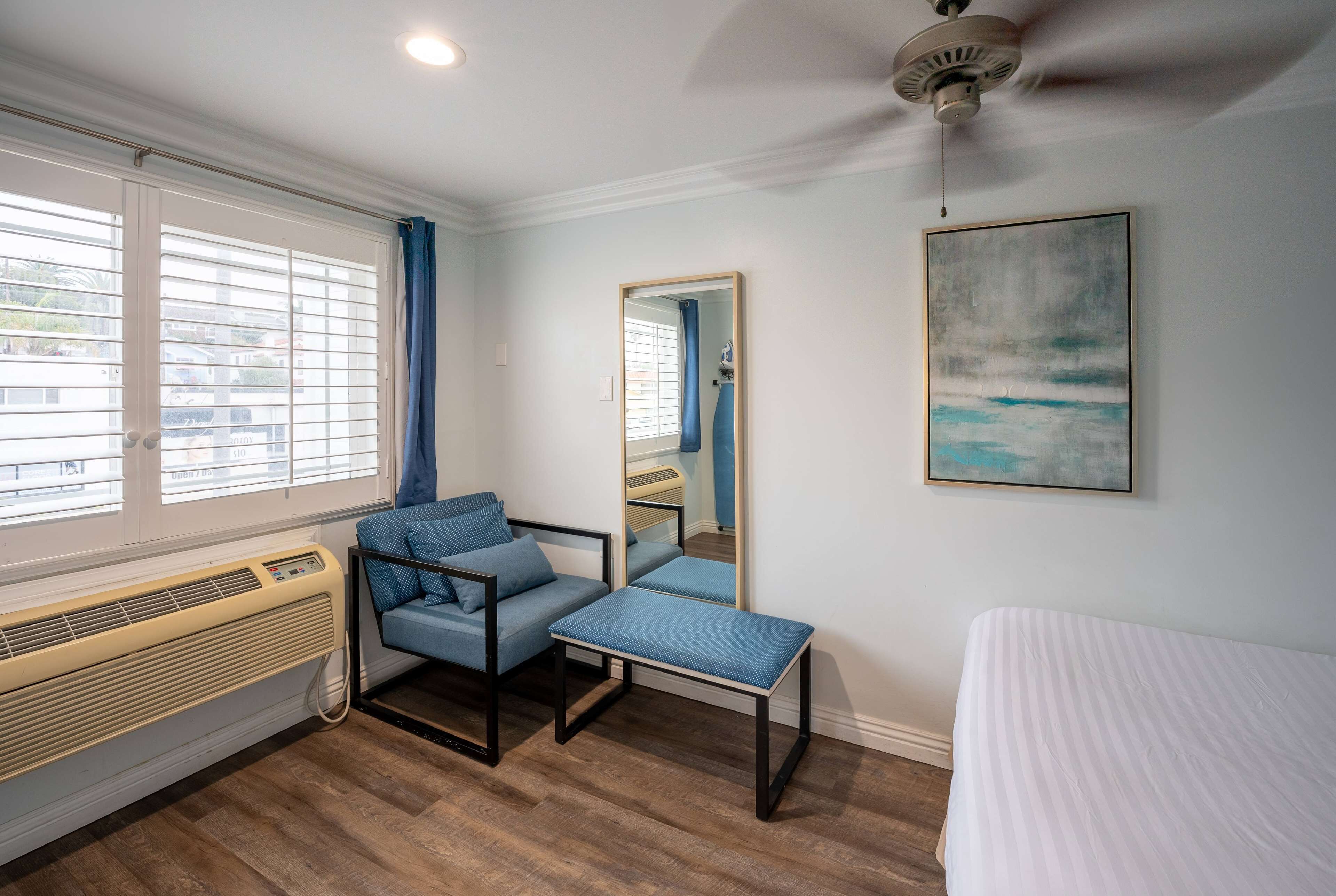 Une chambre à coucher comprenant un lit, une chaise, un banc, un radiateur, un ventilateur de plafond, une fenêtre avec des rideaux et un tableau accroché au mur.