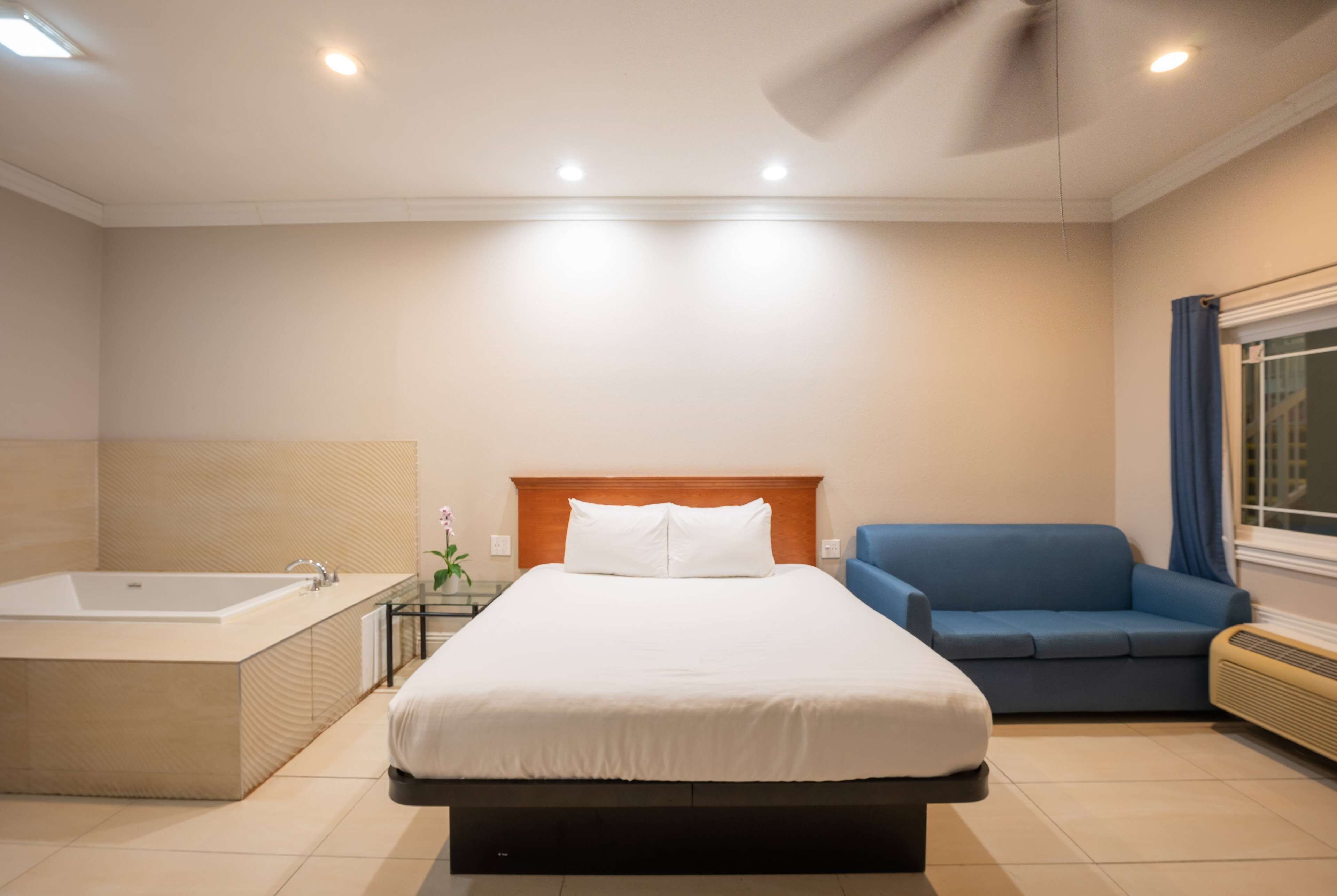 A modern hotel room with a large bed, a separate bathtub, a blue sofa, and a window with curtains.