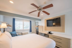 Una habitación de hotel con cama, escritorio, televisor montado en la pared, ventilador de techo y ventana con cortinas.