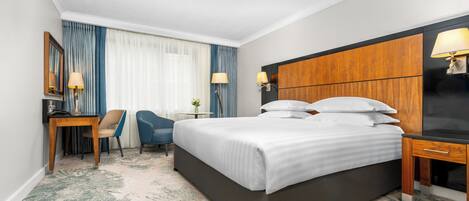 Hotellihuone, jossa on suuri sänky, työpöytä tuolilla, pieni pöytä, jossa on kasvi, ja televisio.