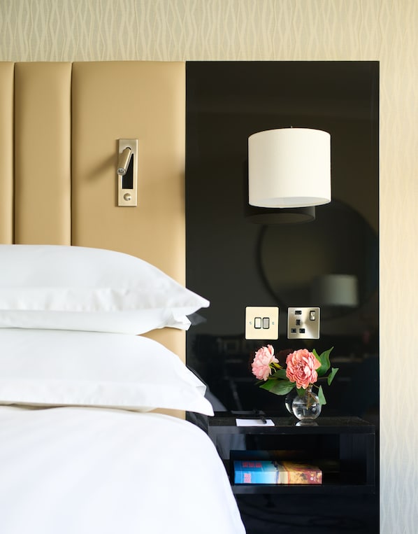 A hotel room with a bed, a nightstand, a lamp, a power outlet, and a vase with flowers.