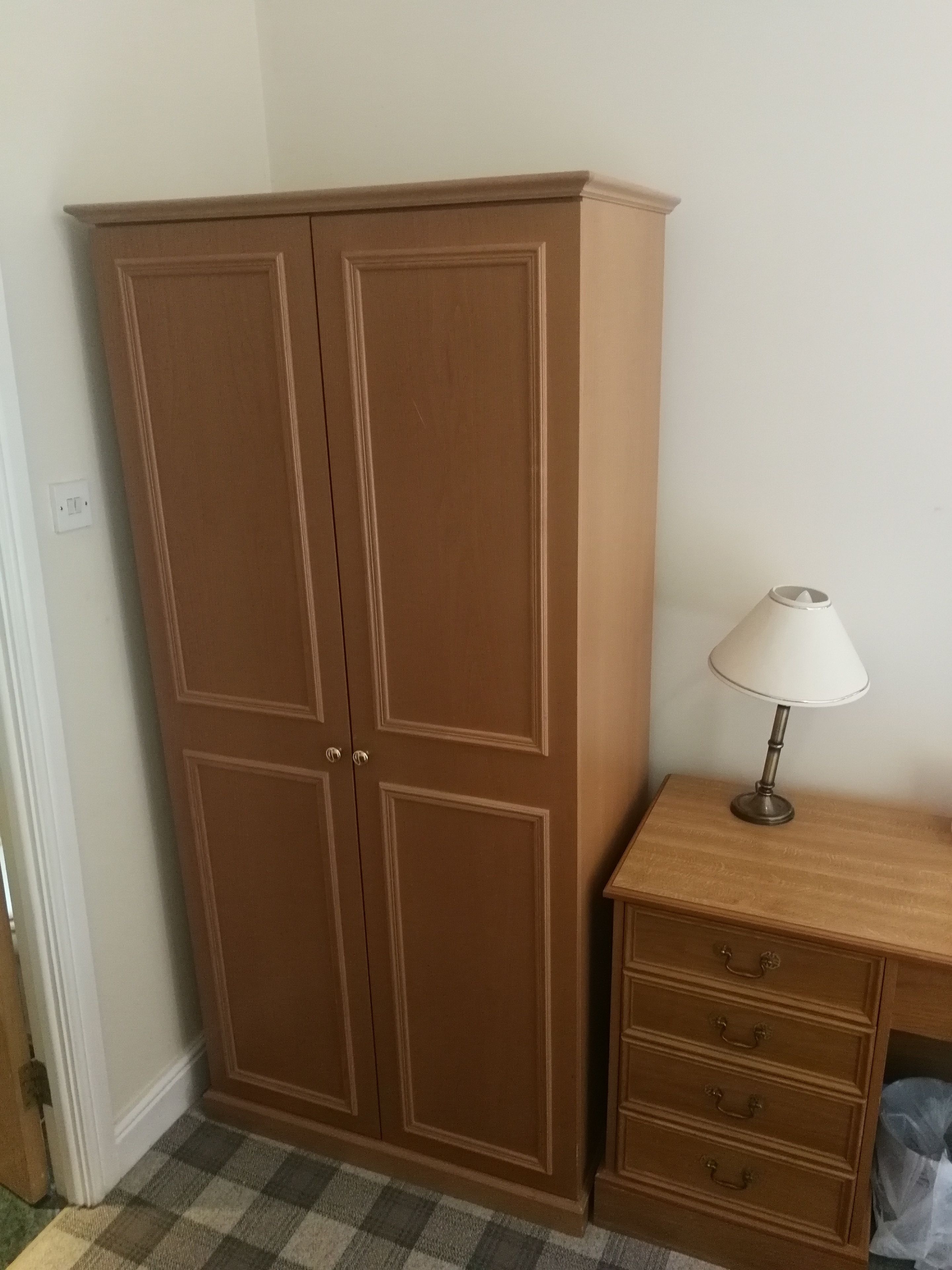 A room with a brown wardrobe, a wooden nightstand with a lamp, and a checkered floor.