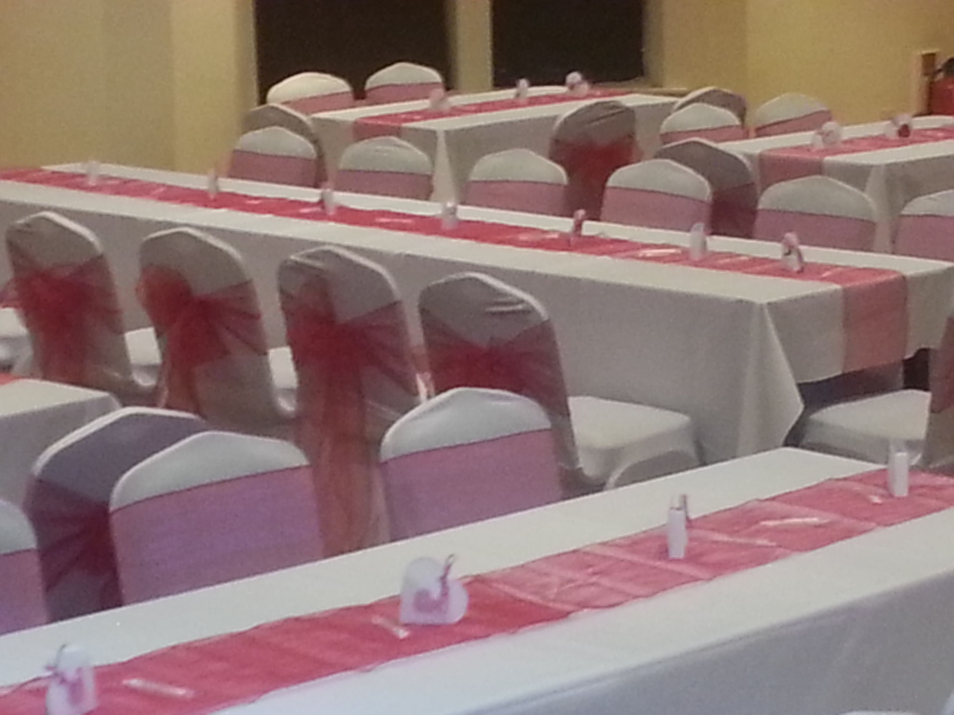 A banquet hall with rows of tables covered in white tablecloths and pink sashes, equipped with white chairs and name placards.