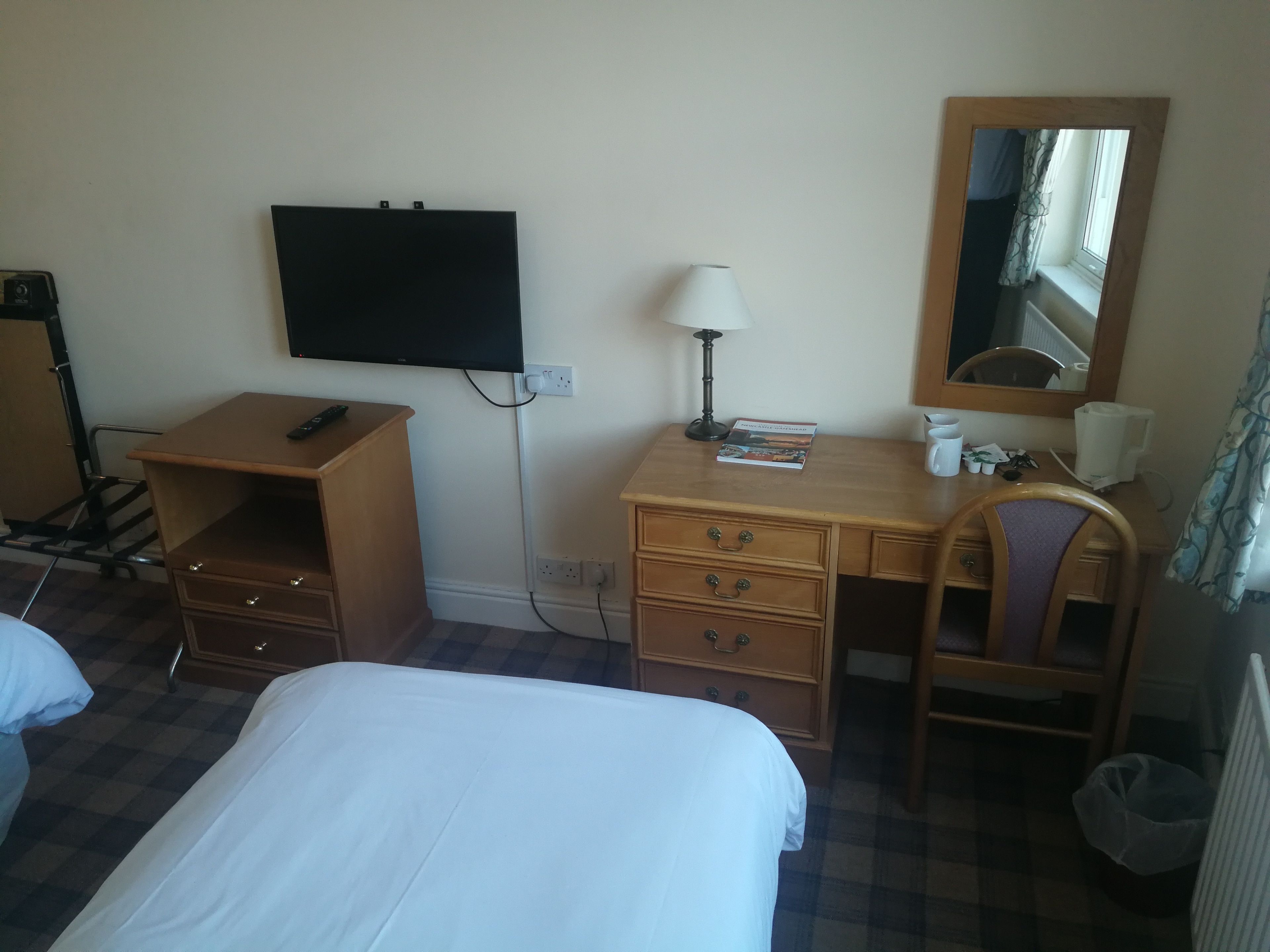 A hotel room with a bed, a desk with drawers, a lamp, a television mounted on the wall, and a mirror.