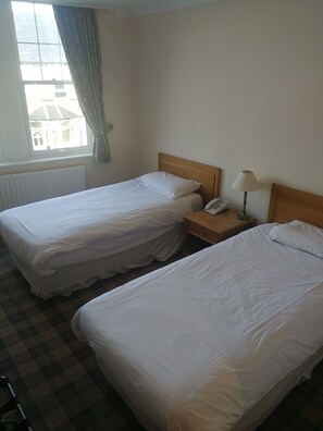 A hotel room with two single beds, a wooden nightstand with a lamp, a telephone, and a window with curtains. - Windsor Hotel (Whitley Bay)