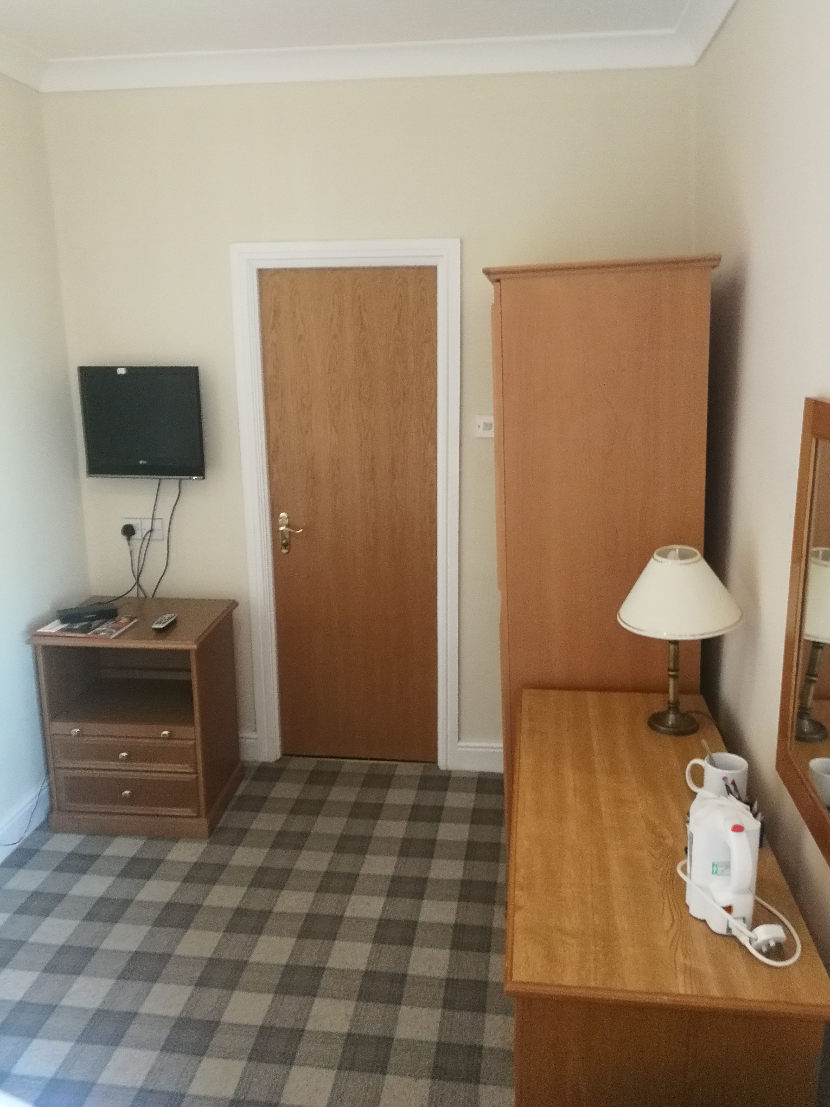 Une chambre d’hôtel avec une armoire en bois, un petit bureau avec une lampe, un téléviseur fixé au mur et un sol à carreaux.