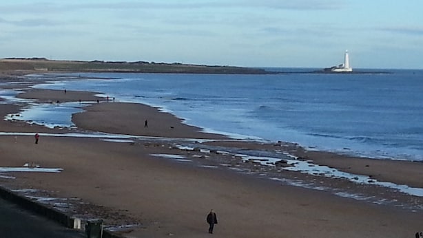 Una playa con un faro a lo lejos, algunas personas caminando y una carretera que corre paralela a la orilla.