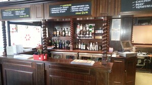 Zona de bar con paneles de madera, una variedad de botellas de licor y una pizarra con un menú que incluye opciones como 'CHARGRILLED STEAKS' y 'BEER'.