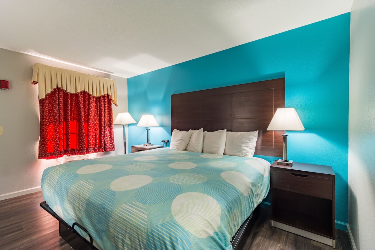 A hotel room with a bed, two bedside tables with lamps, a nightstand, and a window with red curtains.