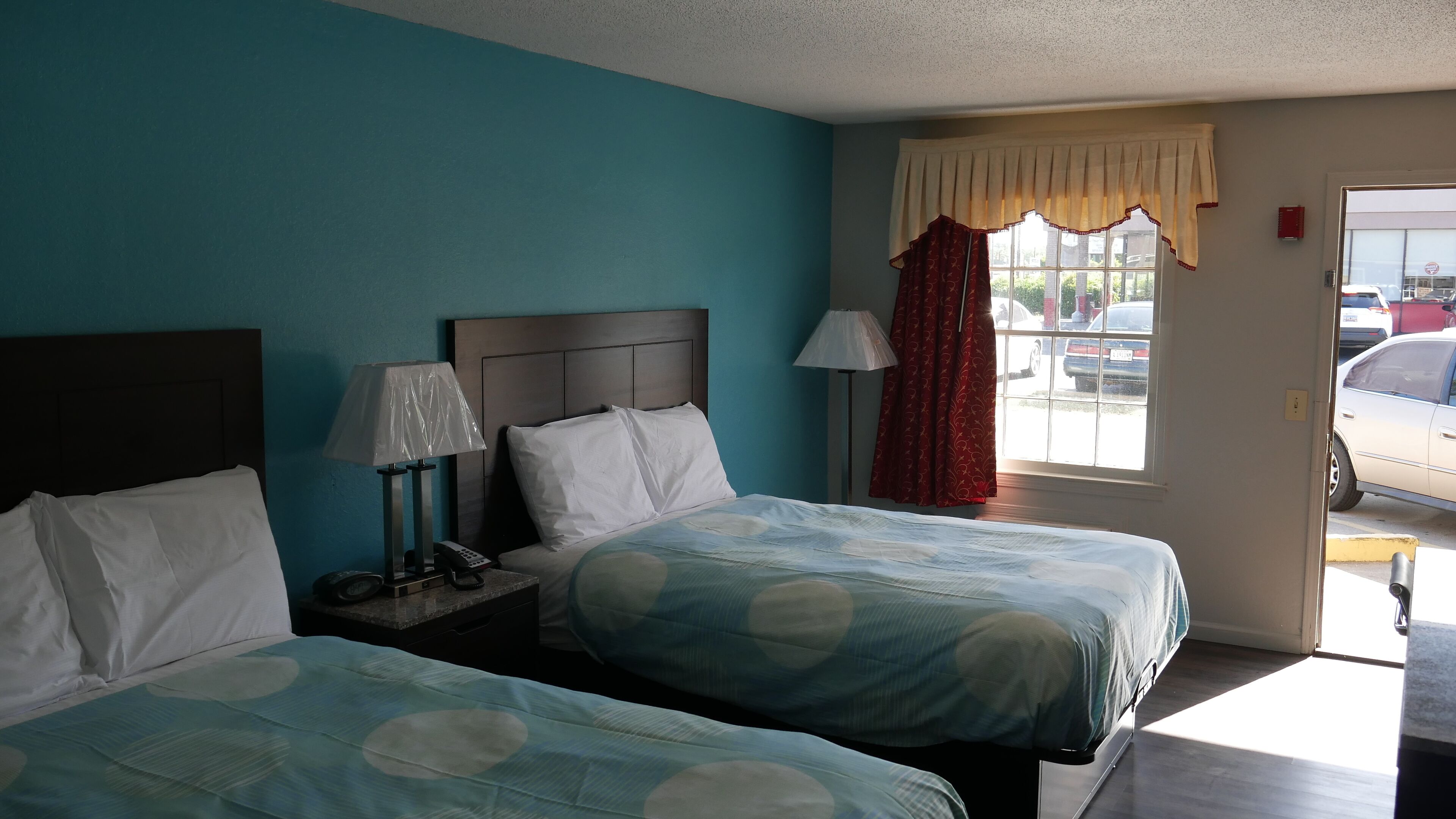 A hotel room with two beds, a nightstand with a lamp and phone, a window with curtains, and a view of parked cars outside.