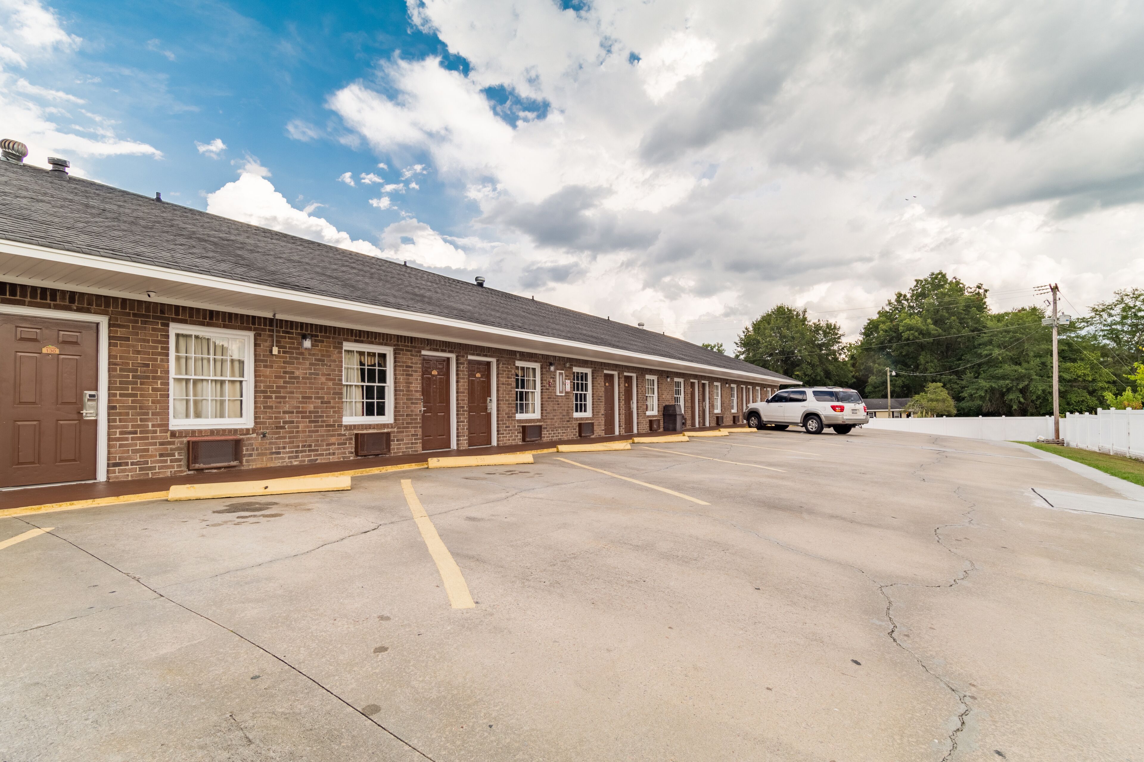 A single-story brick motel with a row of rooms, a parking lot, and a white fence in the background.