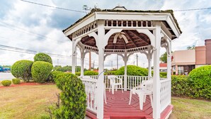A gazebo with a red floor and white railings, surrounded by trimmed bushes and a paved area. - Carolina Lodge (Barnwell)