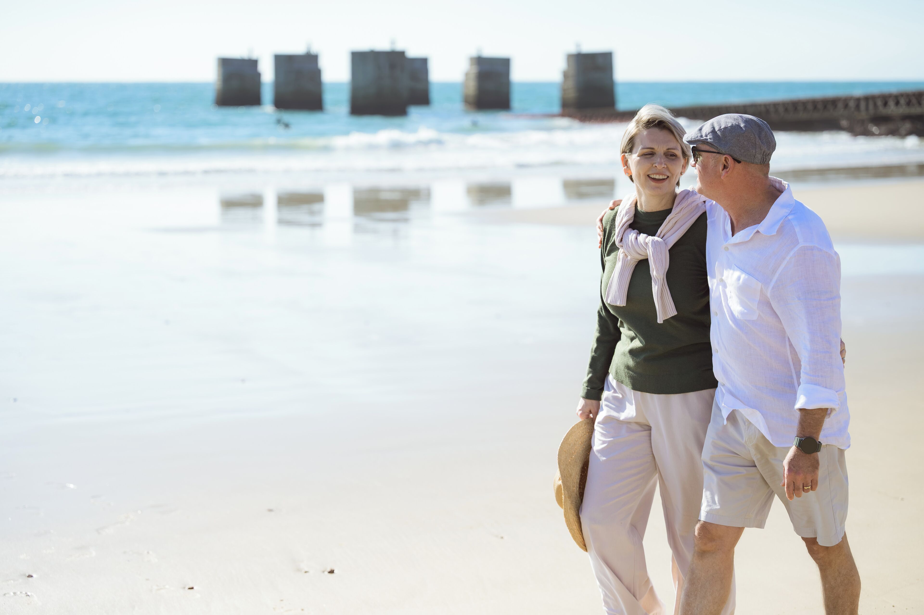A couple walking on a beach with a pier in the background.