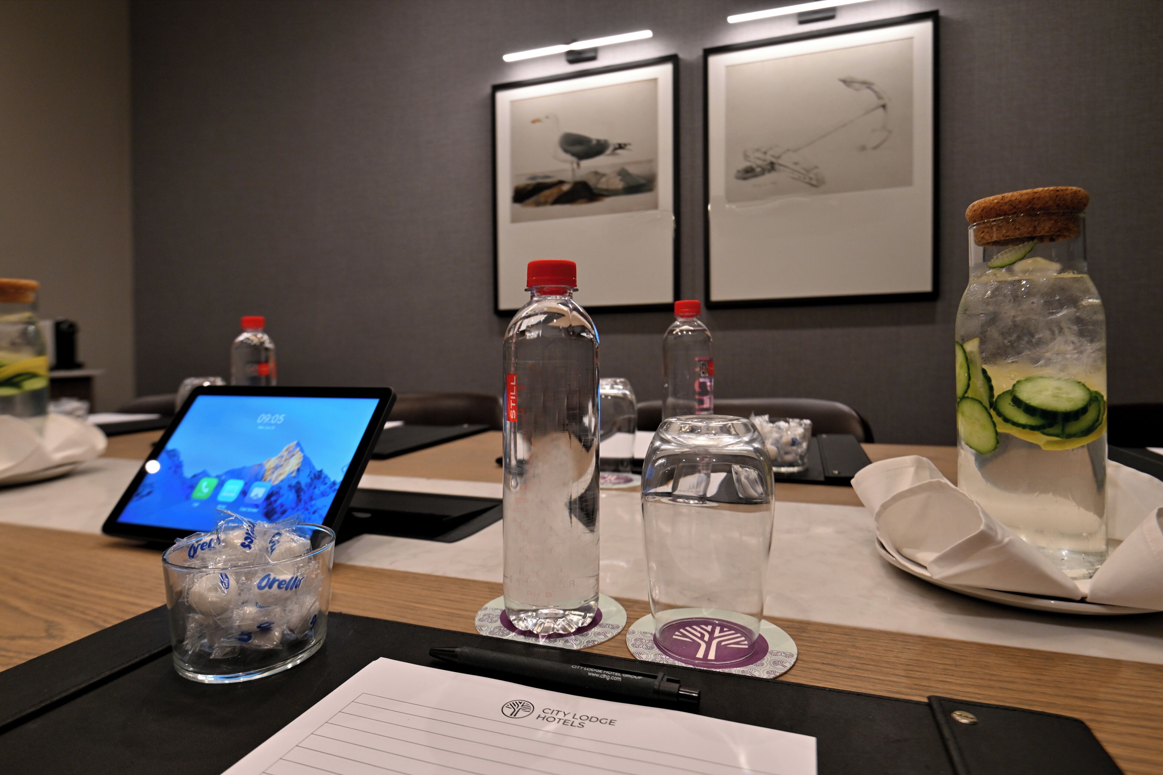 Un salón de conferencias de hotel con una computadora portátil, botellas de agua, un vaso de agua y un posavasos con un logotipo.