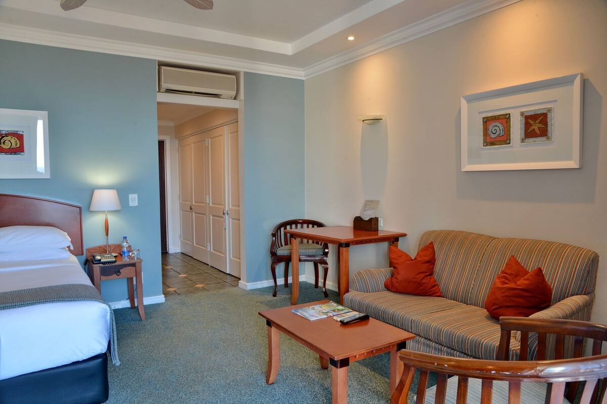 Habitación de hotel con cama, sofá, sillón, una mesita y un escritorio. Hay una puerta que da a otra habitación y un equipo de aire acondicionado en la pared.