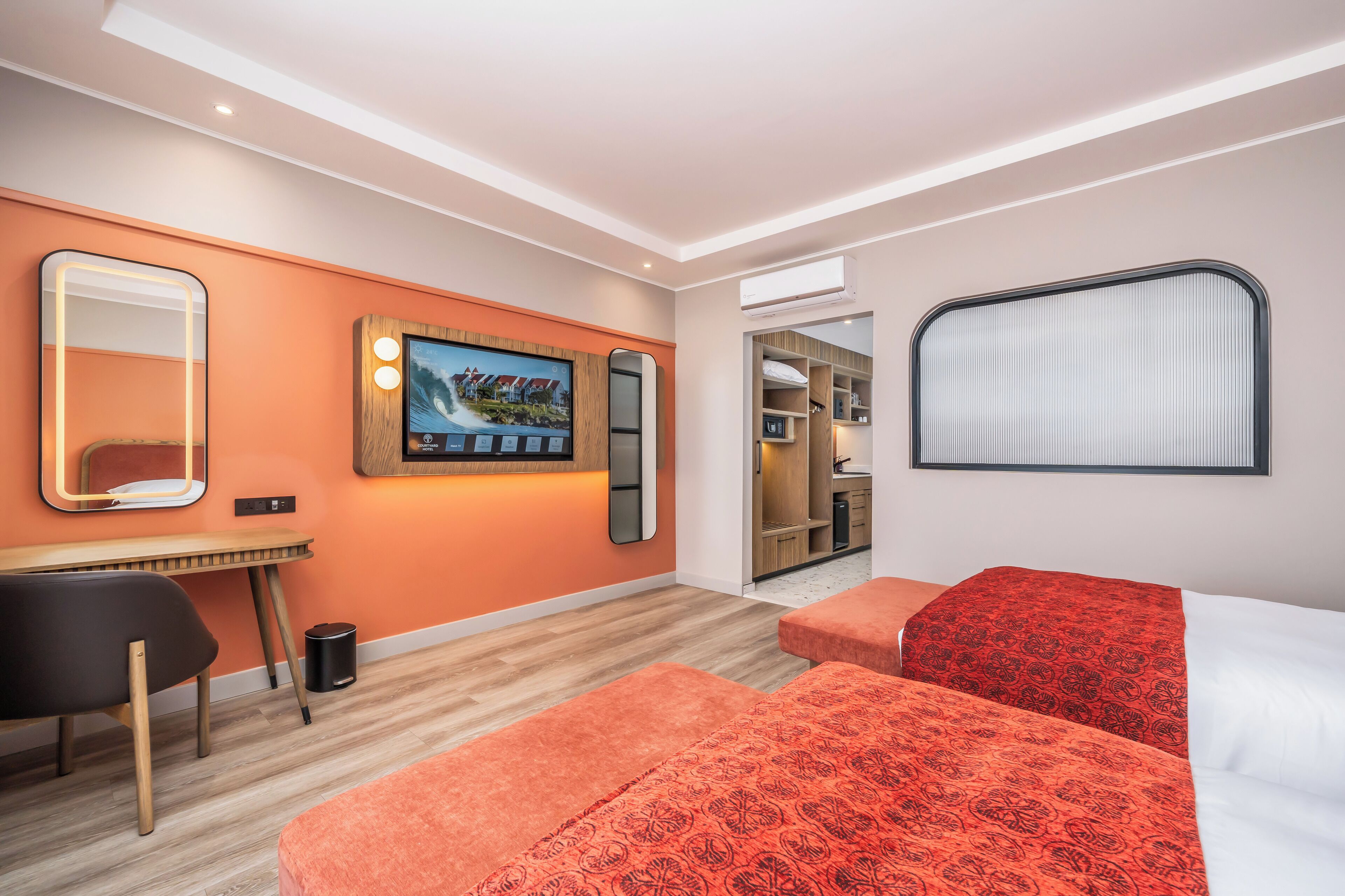 Une chambre d’hôtel moderne avec un bureau en bois, un grand miroir, un téléviseur à écran plat et deux lits avec une literie à motifs rouges.