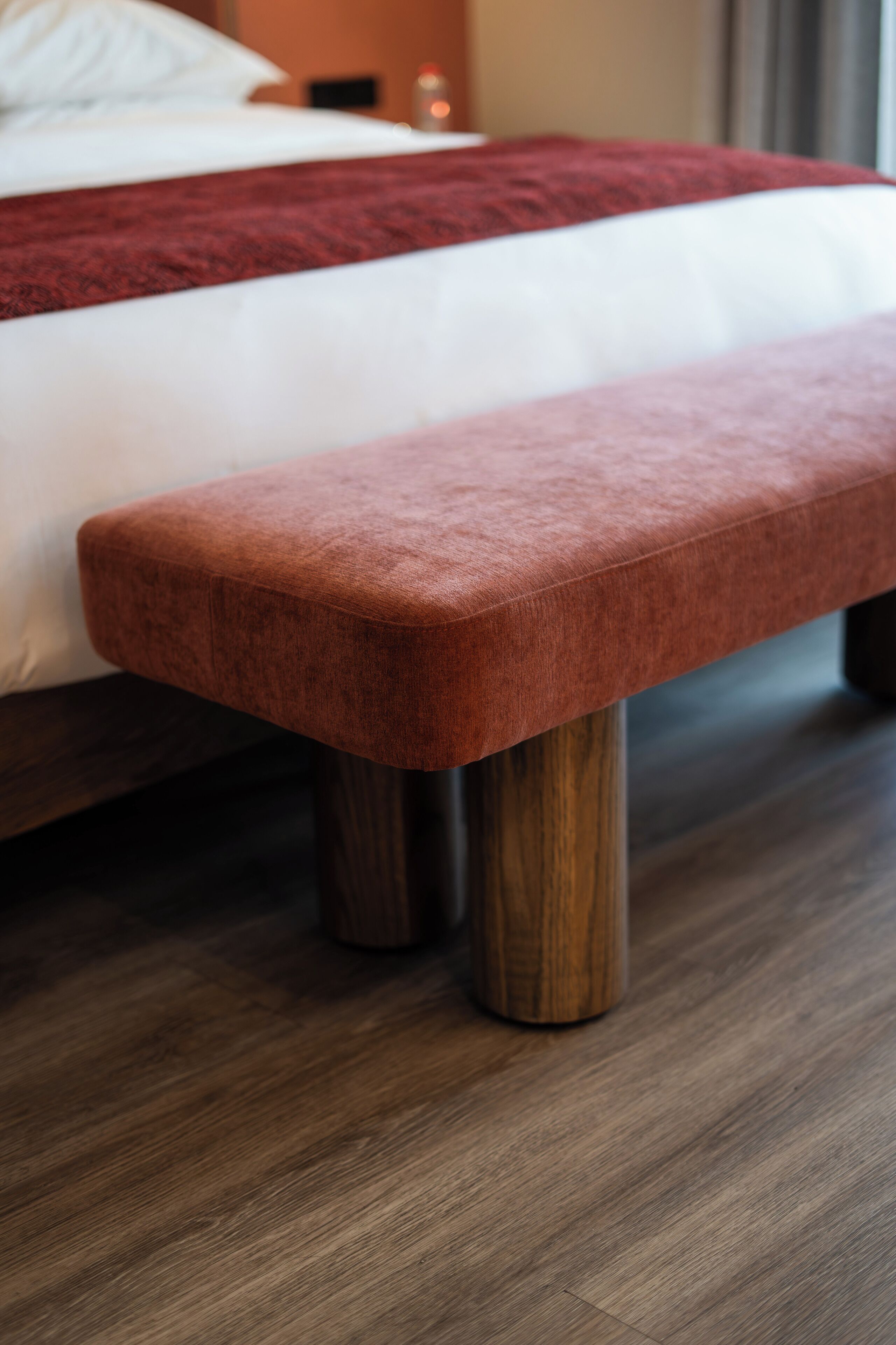 Cama com manta vermelha e um banco de madeira na frente.