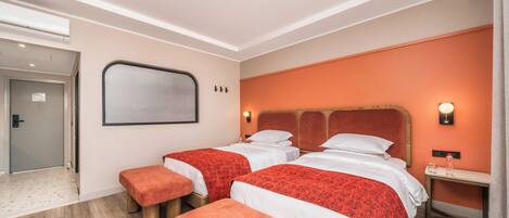 A hotel room with two beds, a bench, a small table, and a wall-mounted light.
