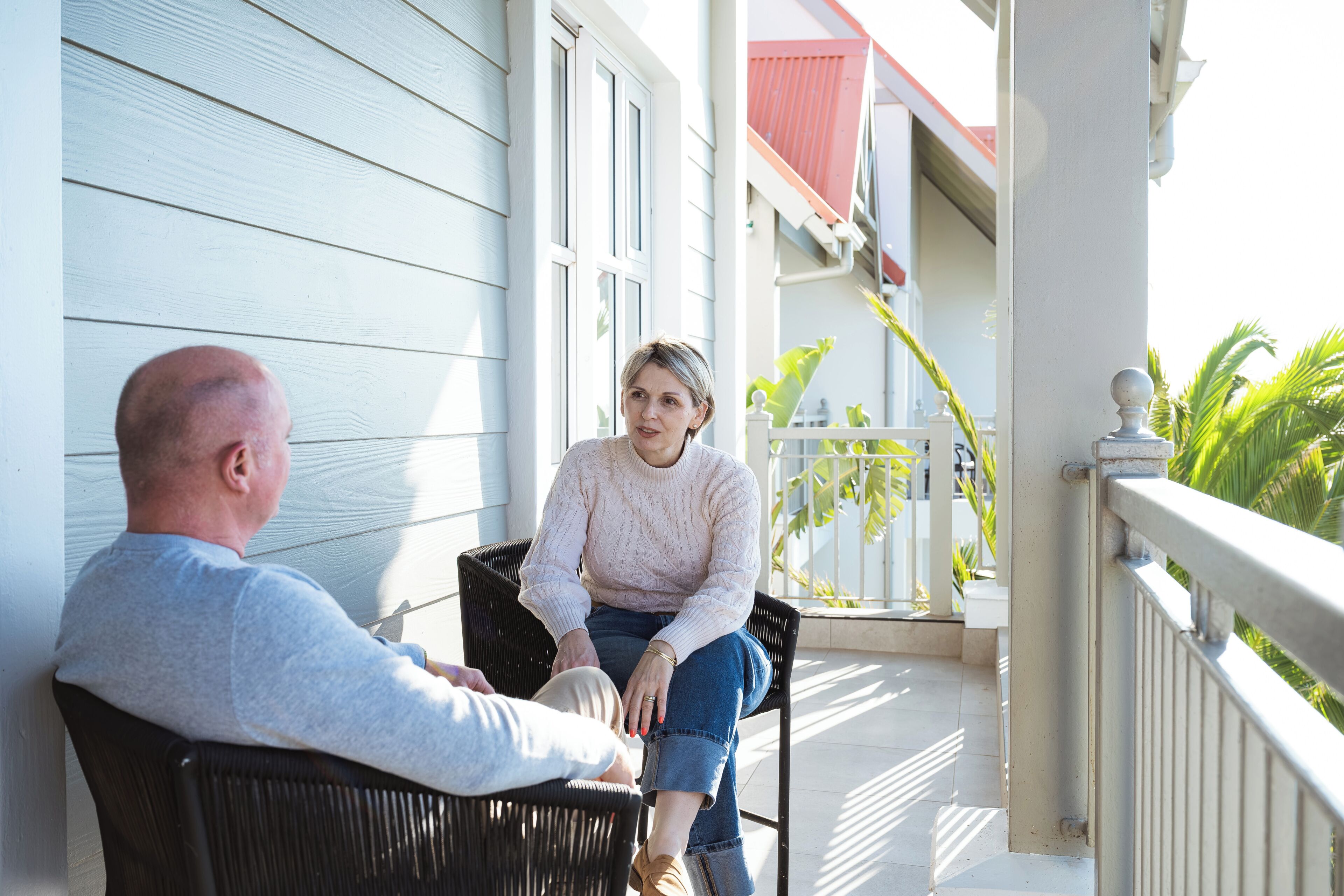 Two people seated on a porch with a railing, a building with a red roof, and a plant in the background.
