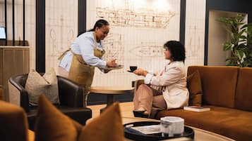 A person serving coffee to another seated individual in a modern interior with a decorative wall and a plant.