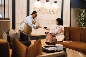 A person serving coffee to another seated individual in a modern interior with a decorative wall and a plant. - Courtyard Hotel Gqeberha (Gqeberha (Port Elizabeth))