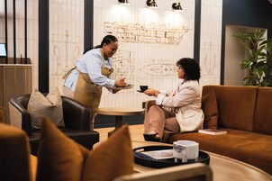 Una persona sirve café a otra sentada en un interior moderno con un muro decorativo y una planta.