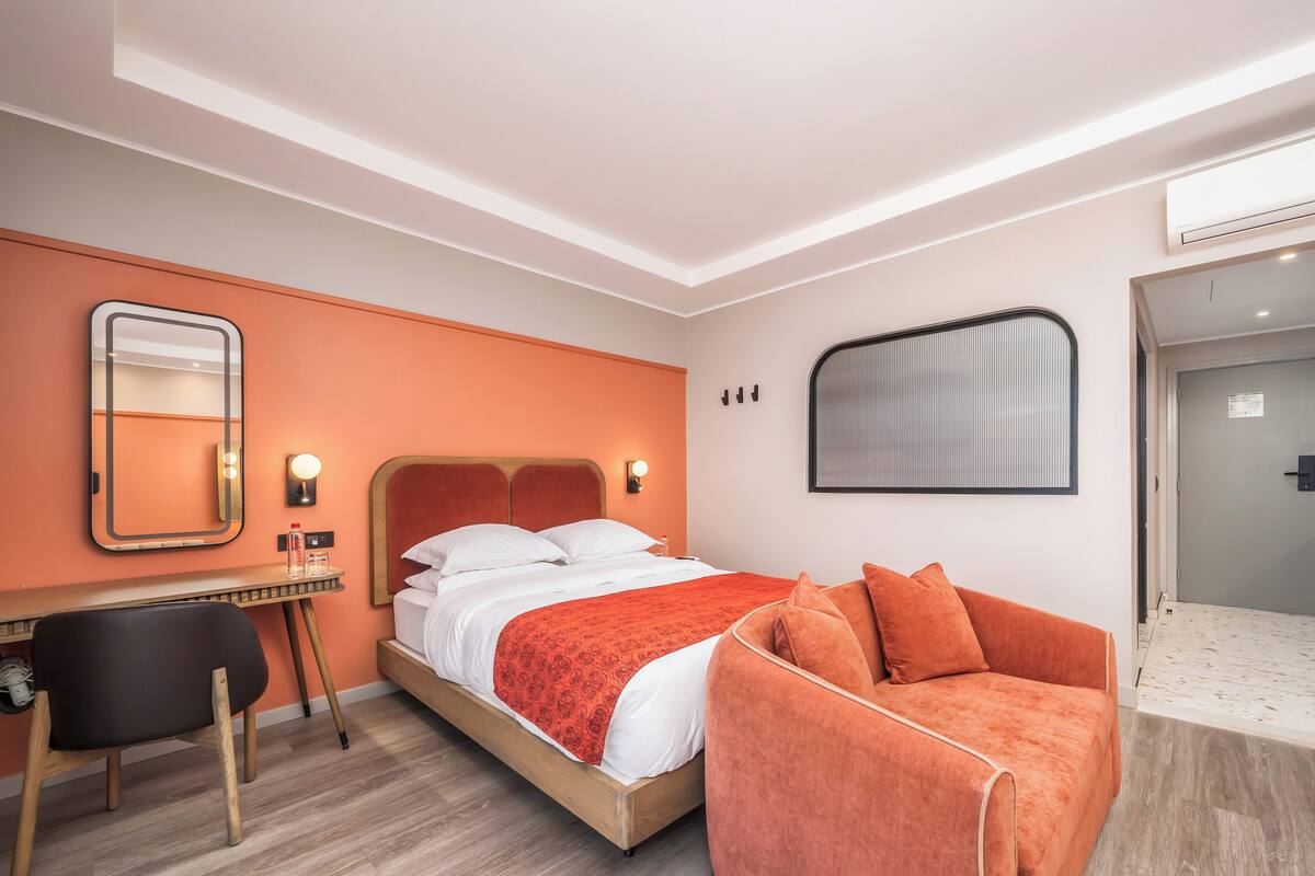 Una habitación de hotel moderna con una cama de madera, paredes de acento naranja, un escritorio con silla y un sofá.