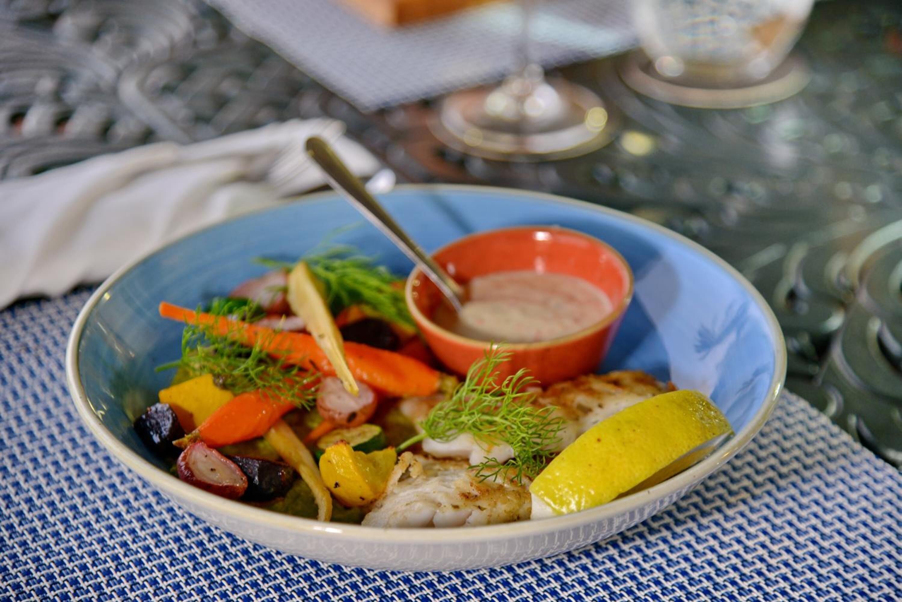 A bowl of fish with vegetables and a side of sauce on a table.