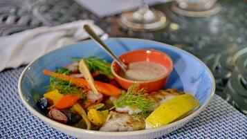 A bowl of fish with vegetables and a side of sauce on a table.