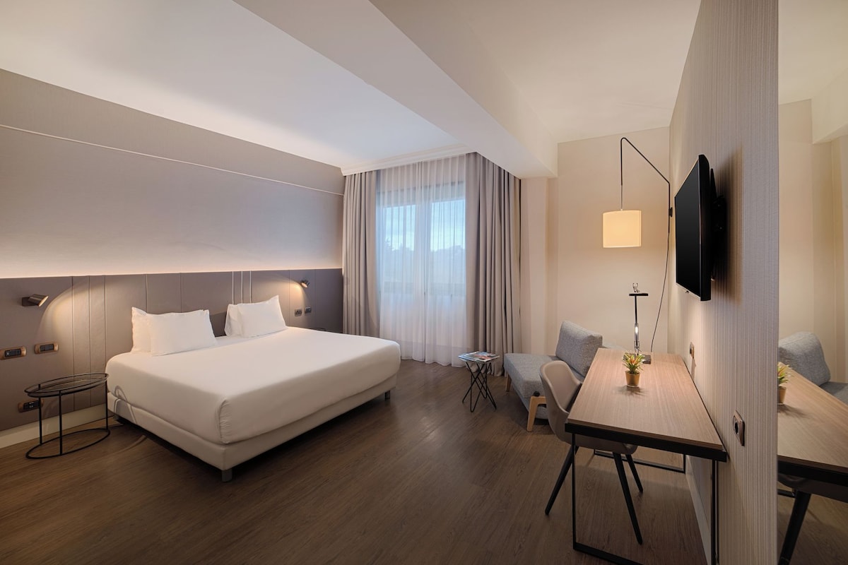 Une chambre d’hôtel moderne, dotée d’un grand lit, d’un bureau, d’une chaise, d’une télévision et d’une fenêtre avec des rideaux.