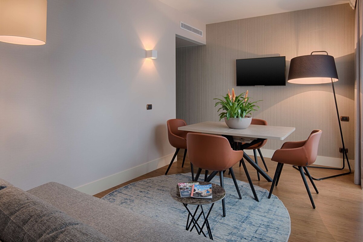 A modern living room with a sofa, a dining table with chairs, a coffee table with magazines, a potted plant, and a TV mounted on the wall.