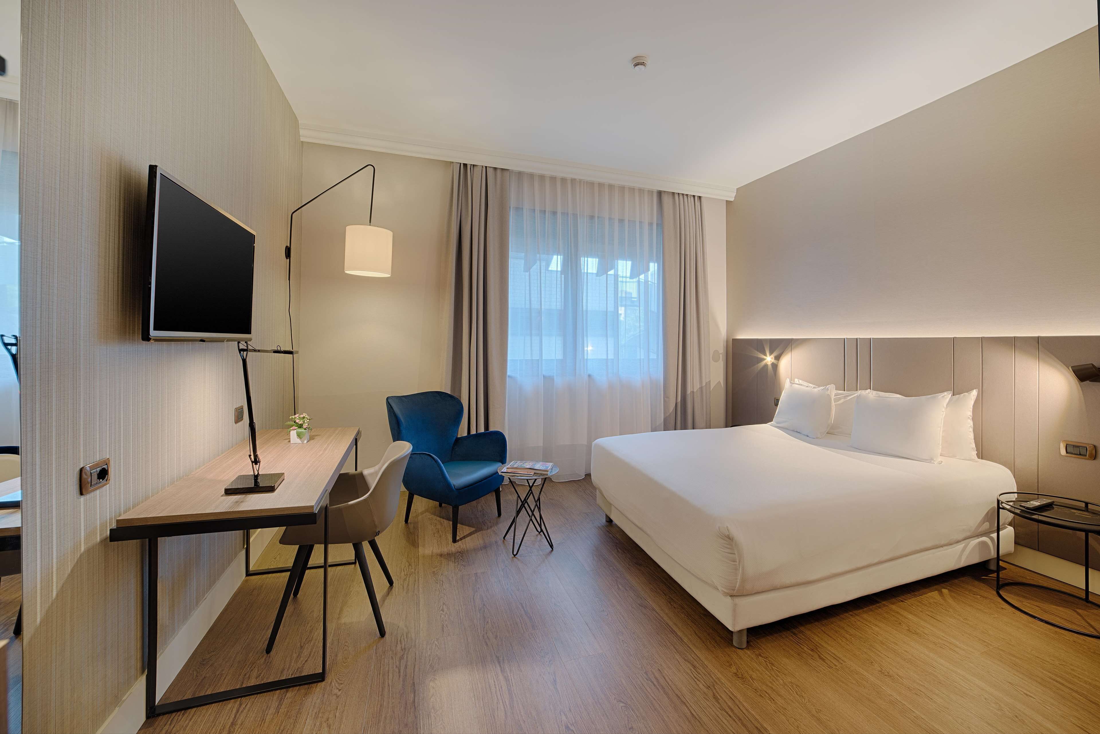 A hotel room with a bed, a desk, a chair, a TV, and a window with curtains.