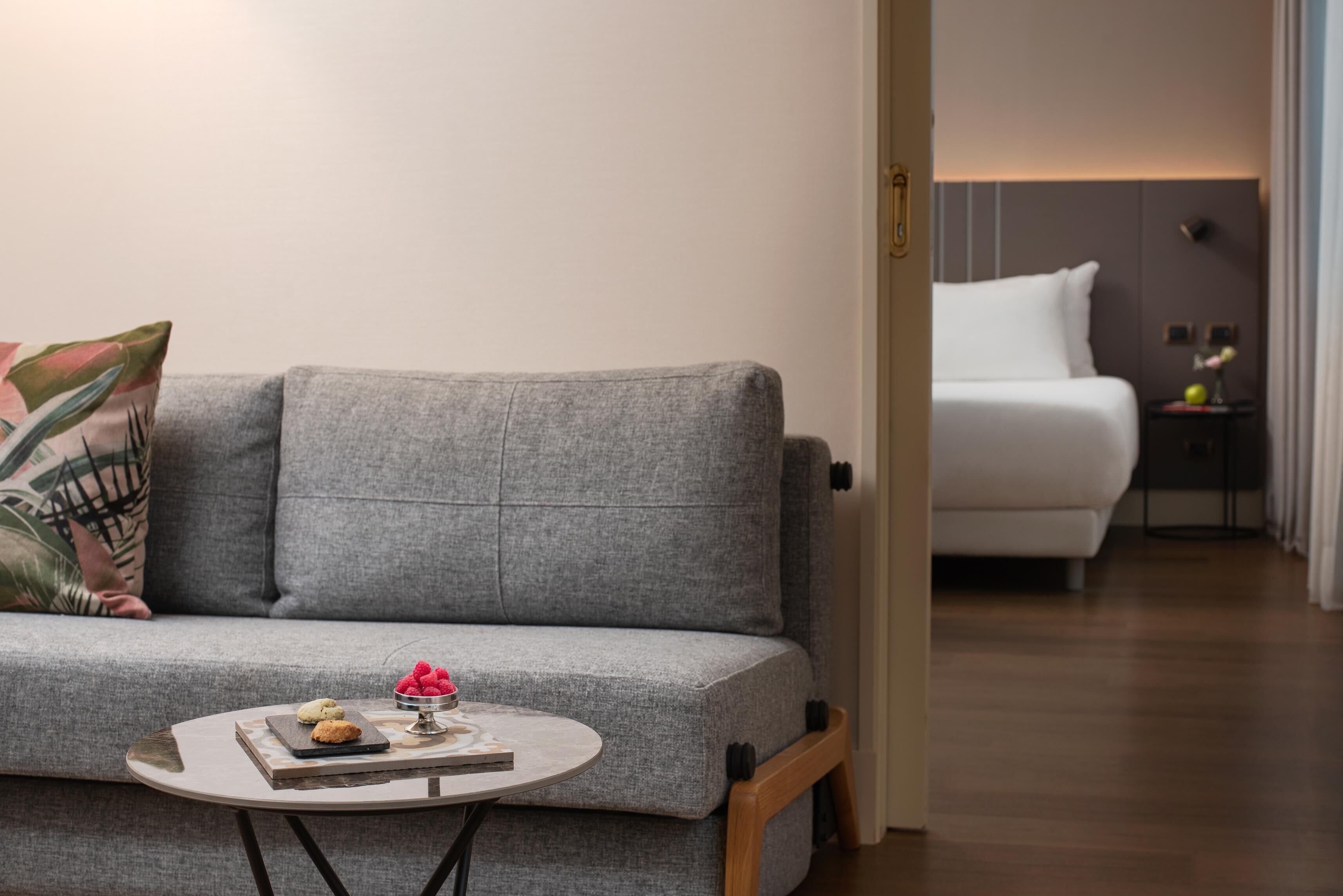 A modern living room with a grey sofa, a small round table with a book and cookies, and a white bed in the background.
