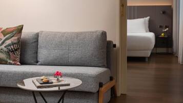 A modern living room with a grey sofa, a small round table with a book and cookies, and a white bed in the background.