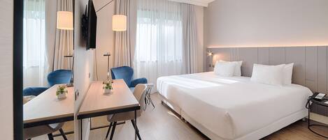 A modern hotel room with a large bed, a desk with a chair, a TV mounted on the wall, and a bedside table with a lamp.