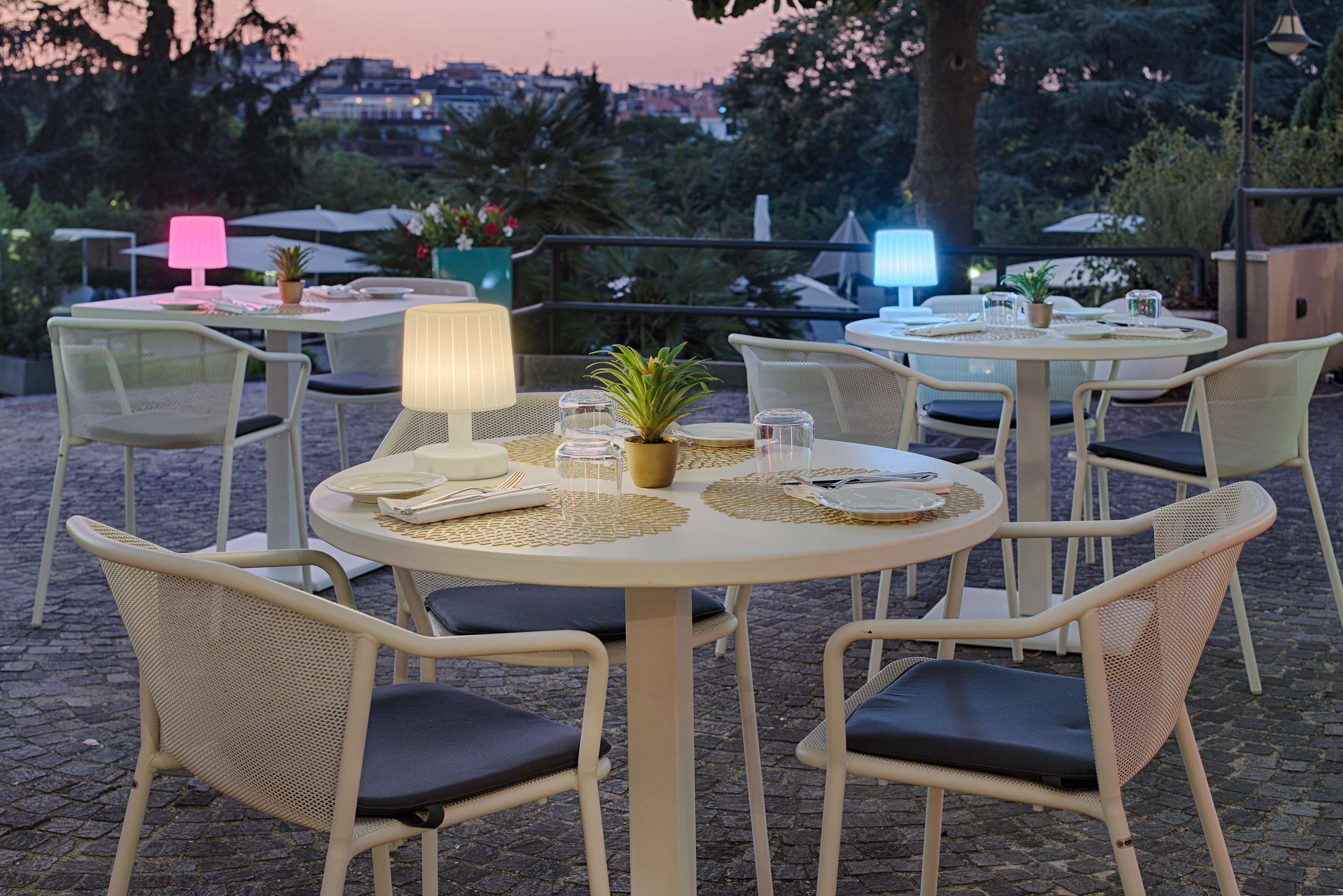 An outdoor dining area with round tables and wicker chairs, equipped with table lamps and potted plants.